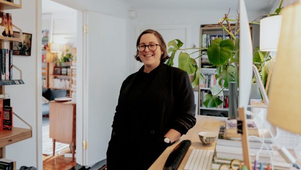 A woman in dark clothing standing at a dlight timber desk in a home office, smiling. Behind her on the desk is a monstera plant in a pot.