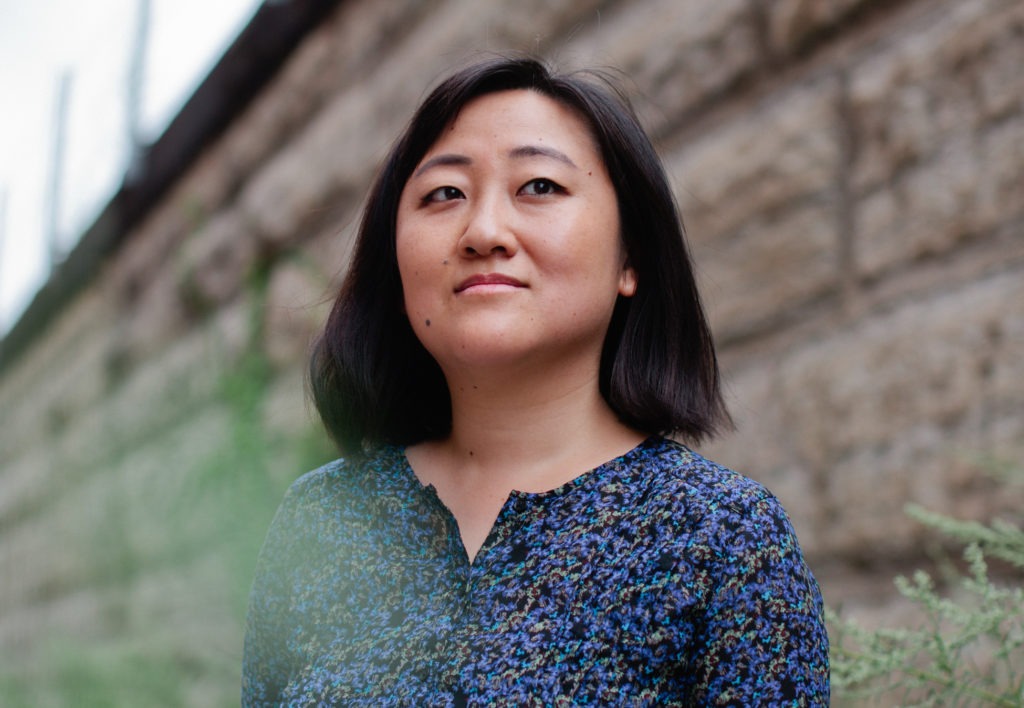 Author Ling Ma wearing a blue dress with a floral pattern, standing among small bushes with a sandstone wall blurred in the background. She has shoulder-length black hair.