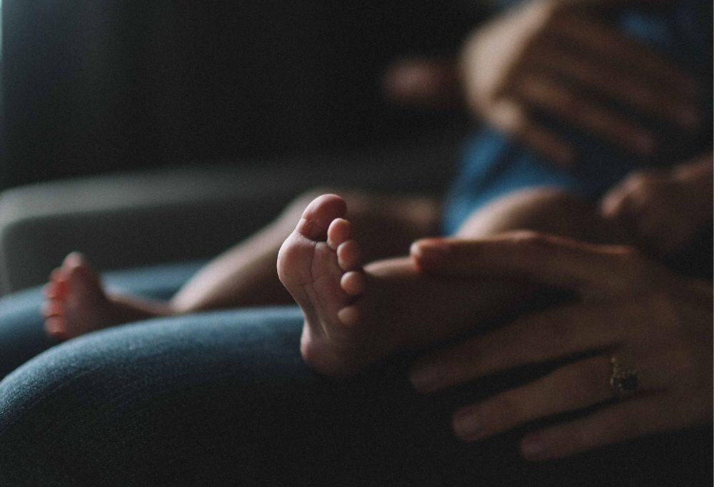 a close-up photo of a baby being held on their parent's lap, focusing on the baby's left foot. The mother and baby are sitting in dim, soft light that casts dark shadows.