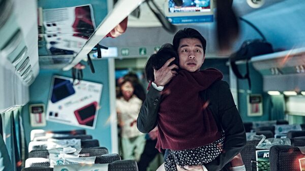 a still from the movie Train to Busan. A man is holding a young girl to his chest and carrying her down the aisle of a train as they are pursued by zombies.