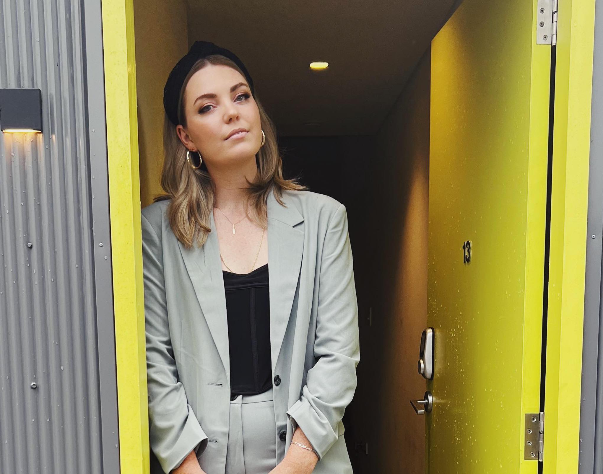 Amy Thunig standing and leaning against a yellow door frame. She is wearing a light grey suit with a black top underneath