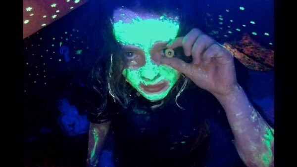 A still from 'We're All Going to the World's Fair'. A grainy image of young girl with her face painted fluorescent green, holding a small round object up to. her eye and leaning directly towards the camera.