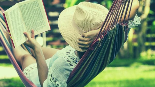 A woman in a white sundress and a large straw hat reading a book in a hammock.
