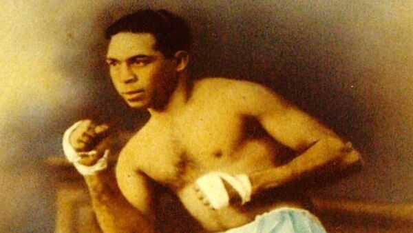 A vintage photograph of An Aboriginal man with short dark hair, wearing white shorts and white strapping around his fists, which are raised in a boxing stance.