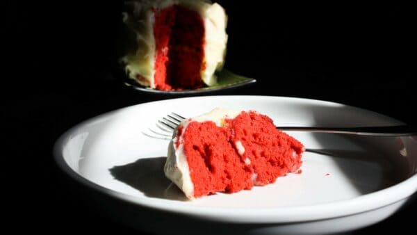 A slice of red velvet cake with white icing, on a white plate next to a metal fork. The rest of the cake sits half in shadow in the background.