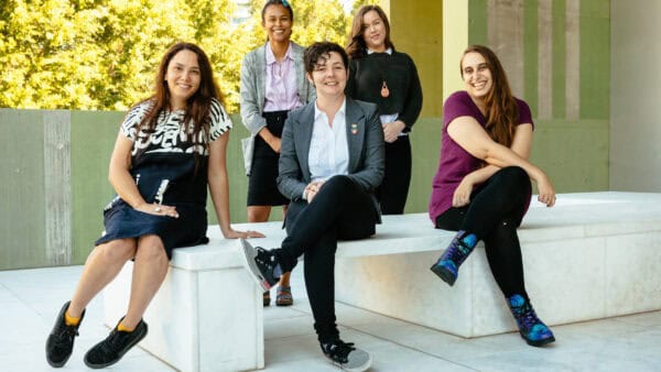 Five women smiling at the camera. Two are standing at the back behind three who are sitting on a light-coloured cement bench.
