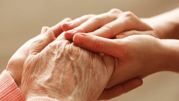A hand belonging to an elderly person, with skin that is papery and wrinkled, is held by the hands of a younger person with smooth youthful skin.
