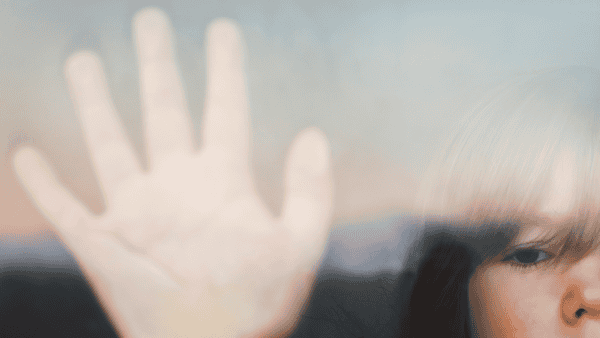 A child stands at a window with their palm against the glass.