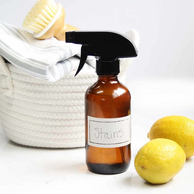 amber glass spray bottle labeled stains next to a white woven laundry basket stacked with cleaning towels and a cleaning brush