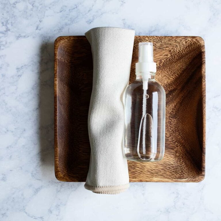small clear glass bottle of DIY stainless steel spray on a wooden tray next to a small microfiber cloth