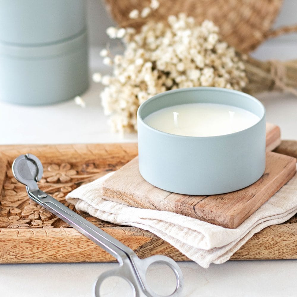 Double wick candle in a sage blue candle tin ontop of wood boards next to a candle wick trimmer.