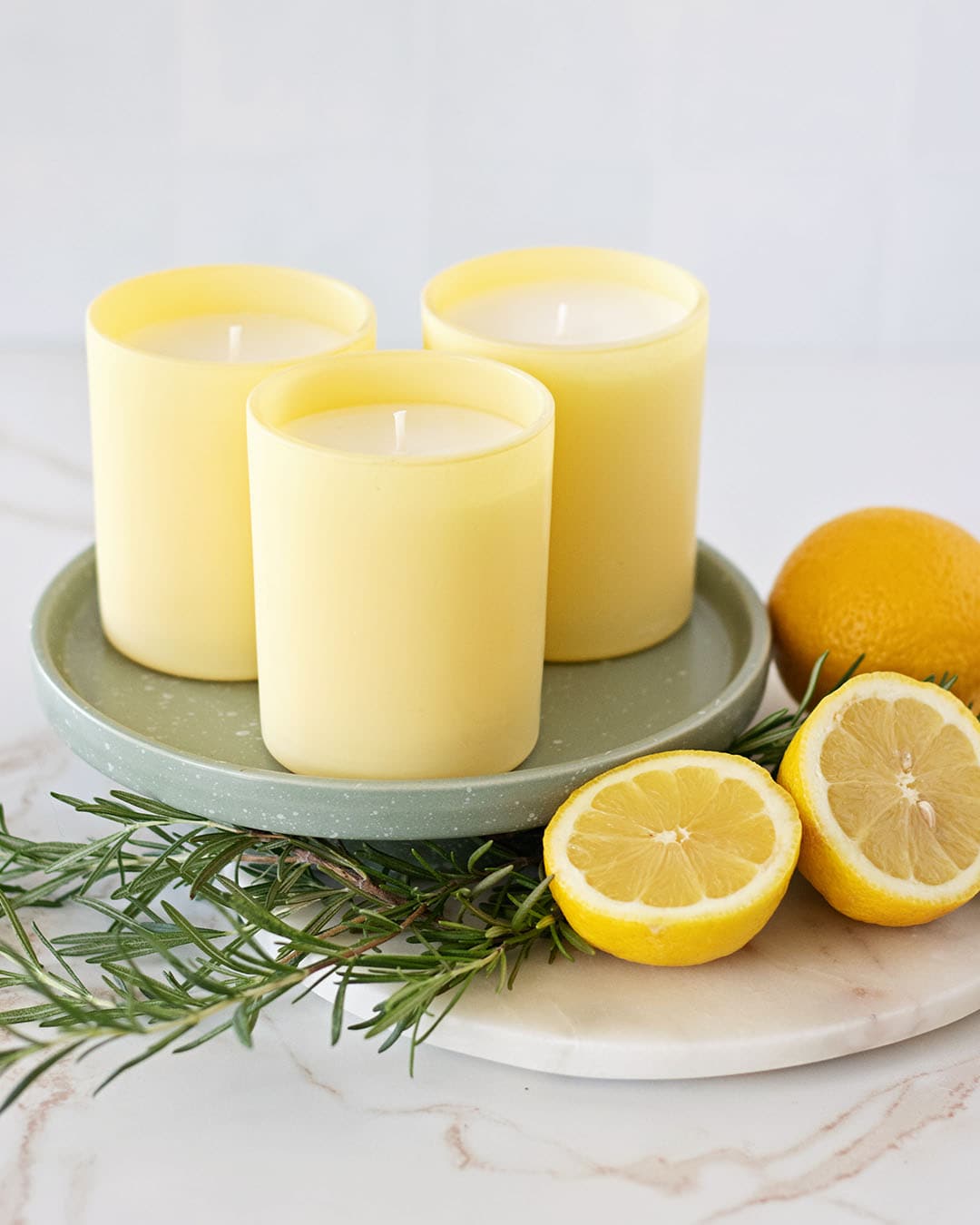 A trio handmade candles in tall sunny yellow candle jars on a sage green tray next to fresh rosemary and sliced lemons halves.