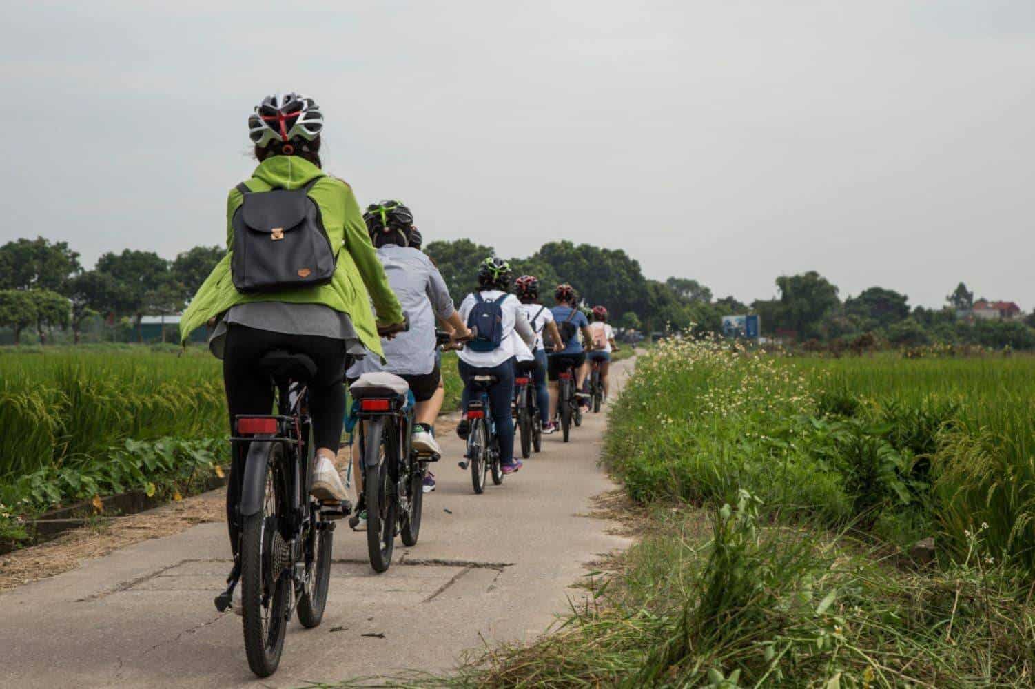 Two cyclists riding along a rural path near Hanoi past green rice paddies and small village houses under an overcast sky