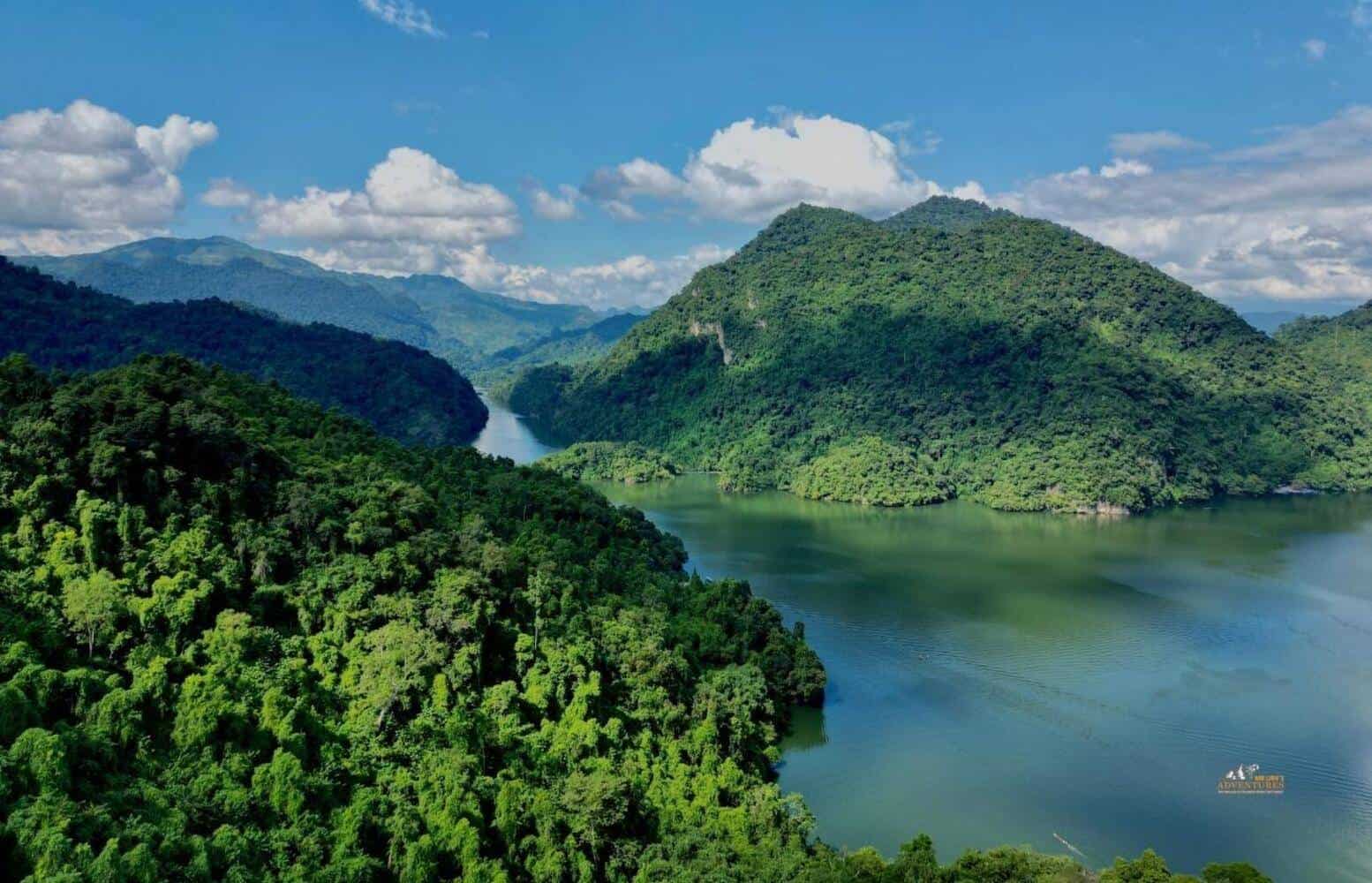 Wide view of Ba Be Lake framed by forested limestone hills with calm green water and a small wooden boat near the shore