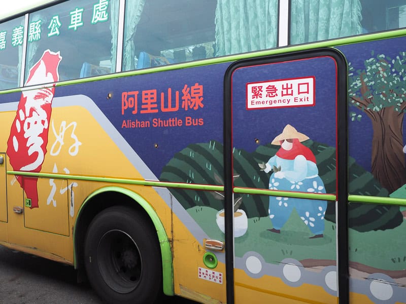 The side of a tour bus that says "Alishan Shuttle Bus" on the side and is painted with a cartoon figure of an old Asian woman and tea fields