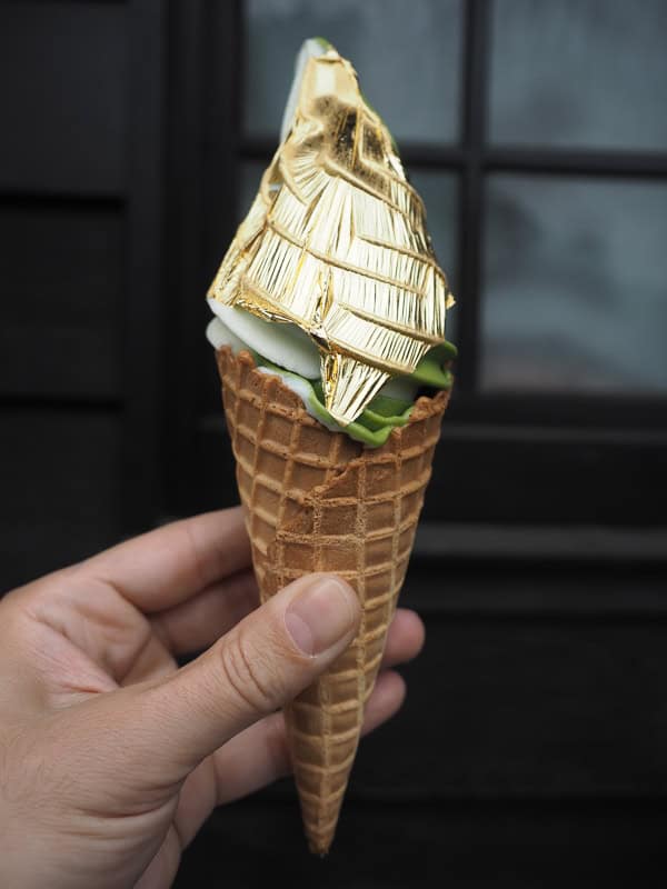 A hand holding up a waffle cone with matcha soft serve ice cream covered with a sheet of thin gold leaf and a black background