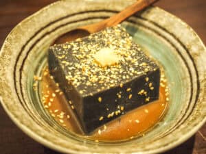 Close up of a rectangular slab of black sesame tofu on a small plate with wooden spoon and garnished with sesame seeds