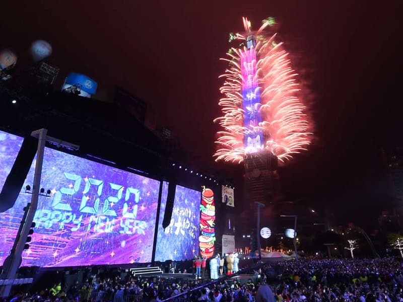 A crowd of people at night in front of stage with LED screen that says 202 Happy New Year and fireworks shooting from the sides of Taipei 101 to the right