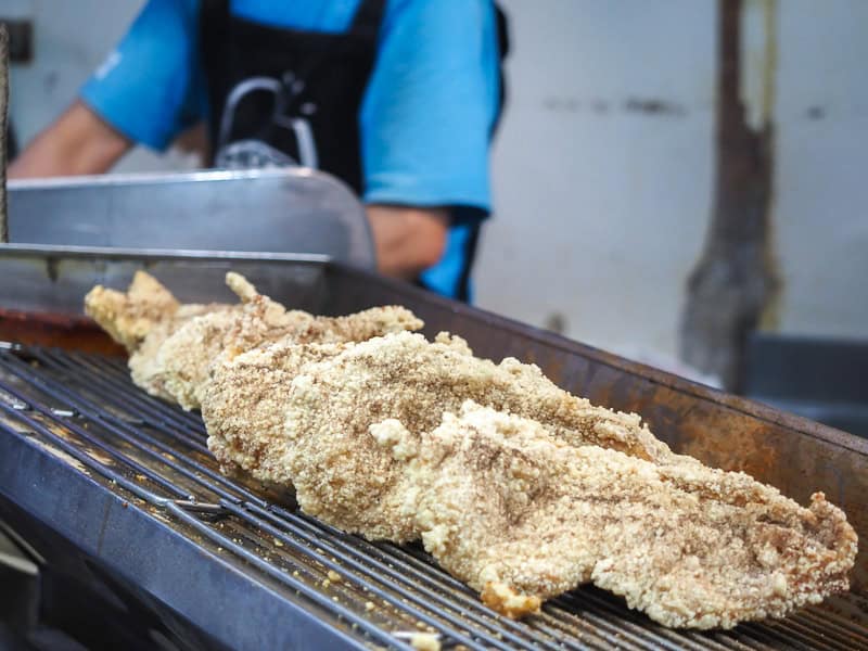 Several huge deep fried breaded chicken cutlets on a metal cooling rack with the midsection of a cook visible behind