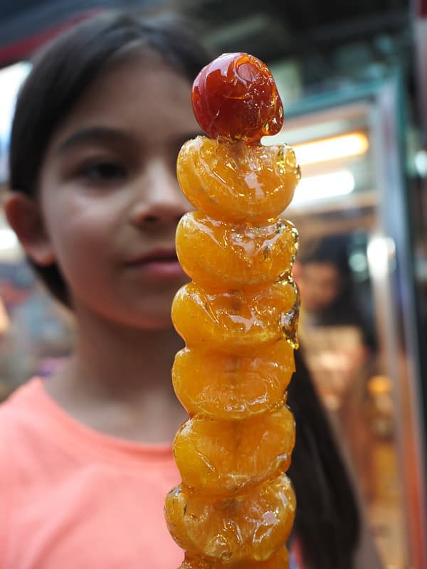 A young girl holding a stick of candied orange and strawberry pieces in front of her face