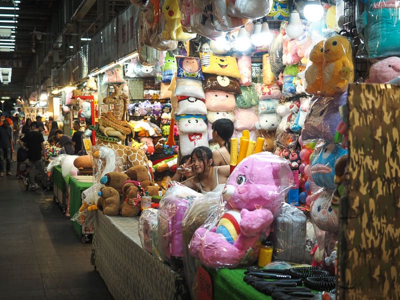 A young Taiwanese woman does the victory sign by her face as she poses for the camera, surrounded by teddy bears and other prizes in a night market games stall
