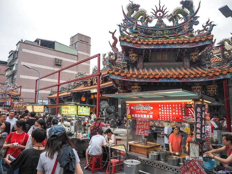 A row of night market food stalls and some shoppers walking by, with a temple behind the stalls