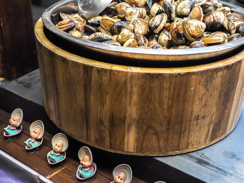 A big wooden tub filled with small clams, and a row of cute characters inside clam shells as decorations on the table in front of it