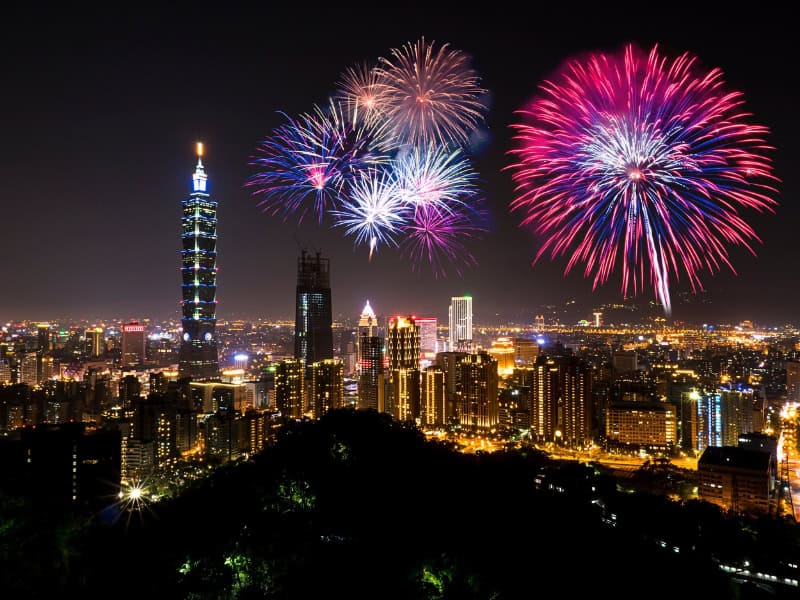 Fireworks exploding above Taipei City at night, shot from high up, with Taipei 101 to the left and a dark hill at the bottom