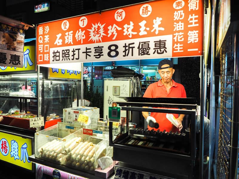 A food vendor wearing red shirt, grilling some corn on the cobs, and with a red and white sign above him