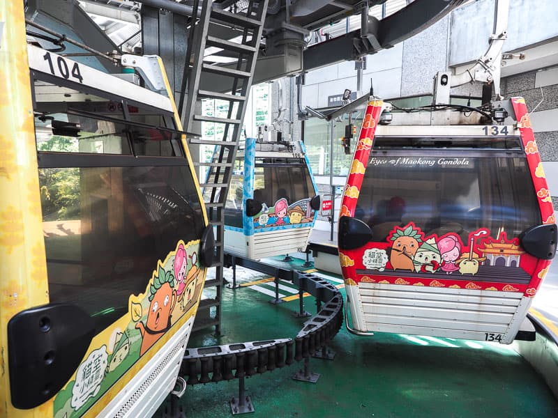 Three cable cars (one yellow, one red, and one blue) curving around inside a gondola station boarding area, with cute cartoon illustrations on their sides