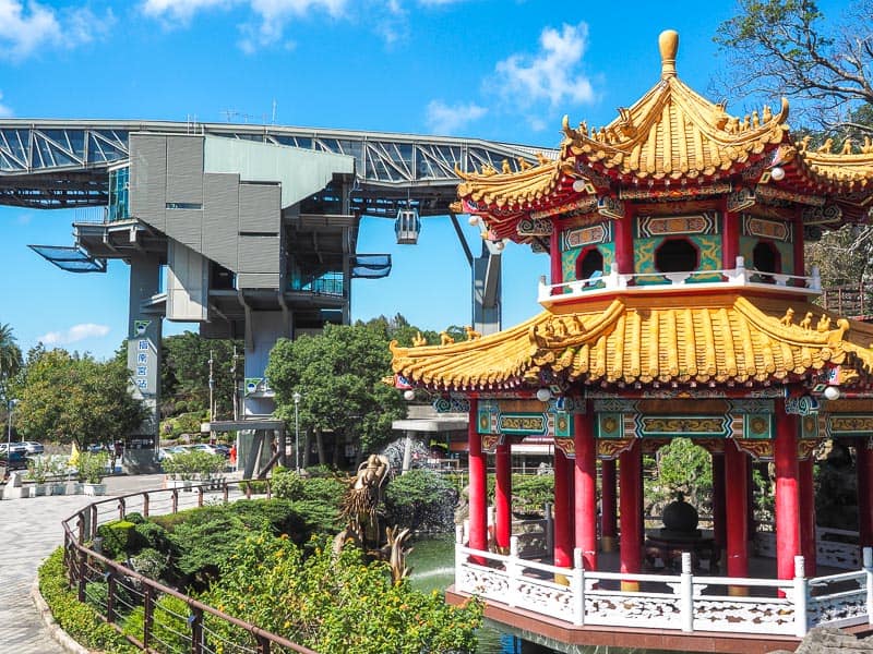 An orange and red Taoist shrine in a pond in the foreground, with tall gondola station tower in background and one gondola car arriving at it