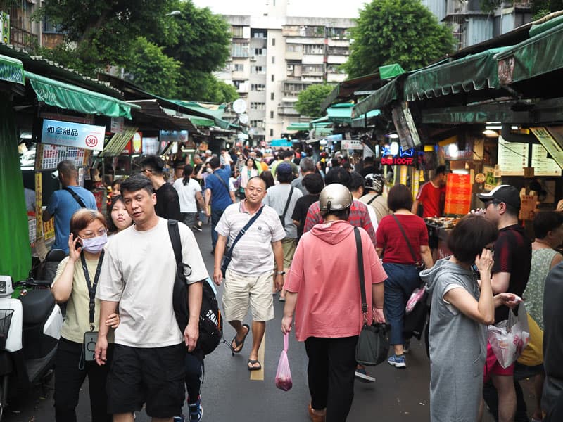 A moderately dense crowd of people walking between food stalls on either side of a narrow street