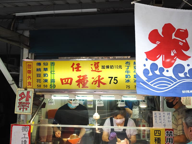 A Taiwanese dessert food stall with yellow sign and menu above and a hanging white and blue poster with Mandarin character 冰 in red on it