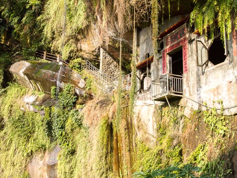 A small temple built into a cliff with all kinds of vegetation hanging from it and small stream pouring down