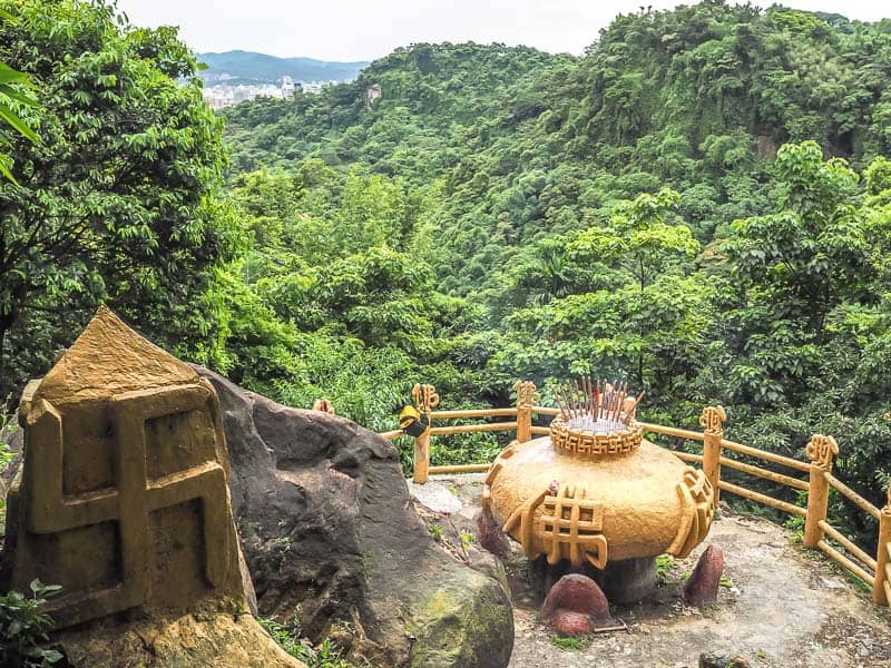 A large golden incense cauldron on a stone lookout platform overlooking forested hills