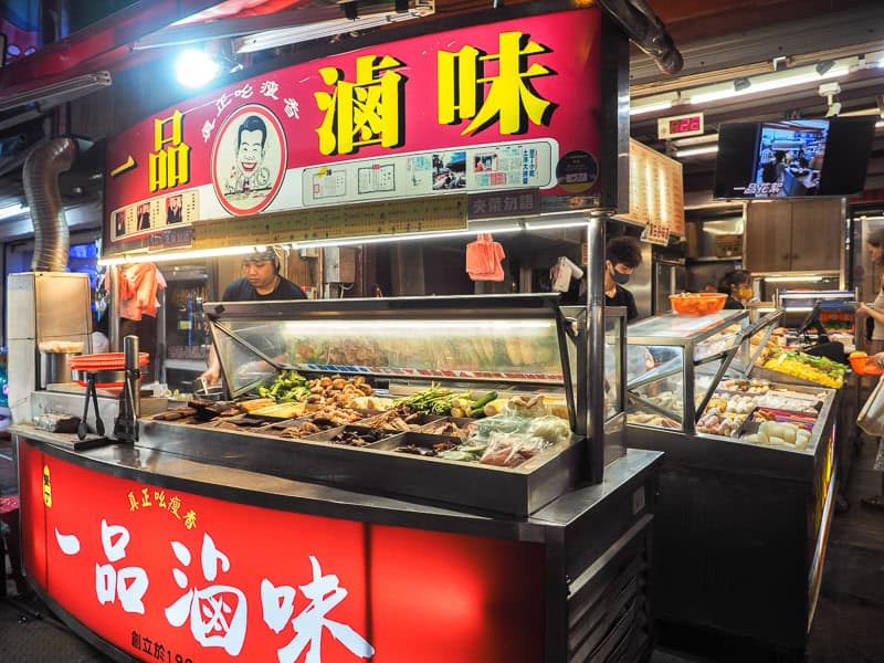 A food stall with red signs, Mandarin name in yellow, various food items on display, and a couple staff working behind it