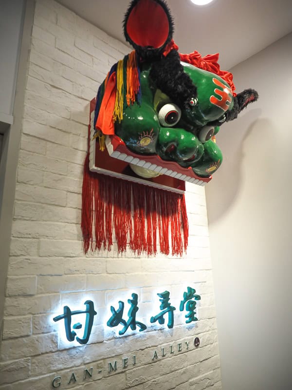 A white brick wall at the top of a staircase in a breakfast shop, with the shop's name on the wall, and a large green and red traditional Chinese lion head above it