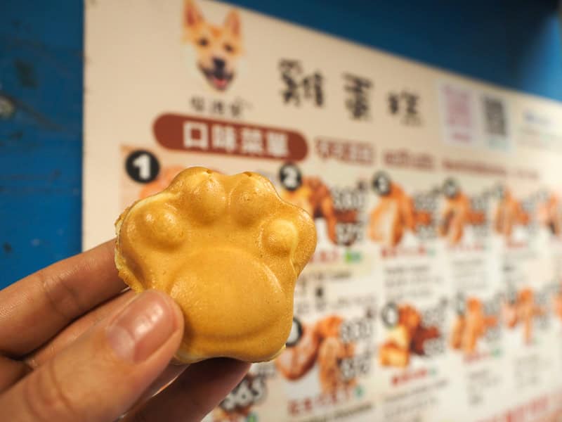 A hand holding up a little cake shaped like a dog's paw, with a menu board on the wall with cute corgi picture behind it