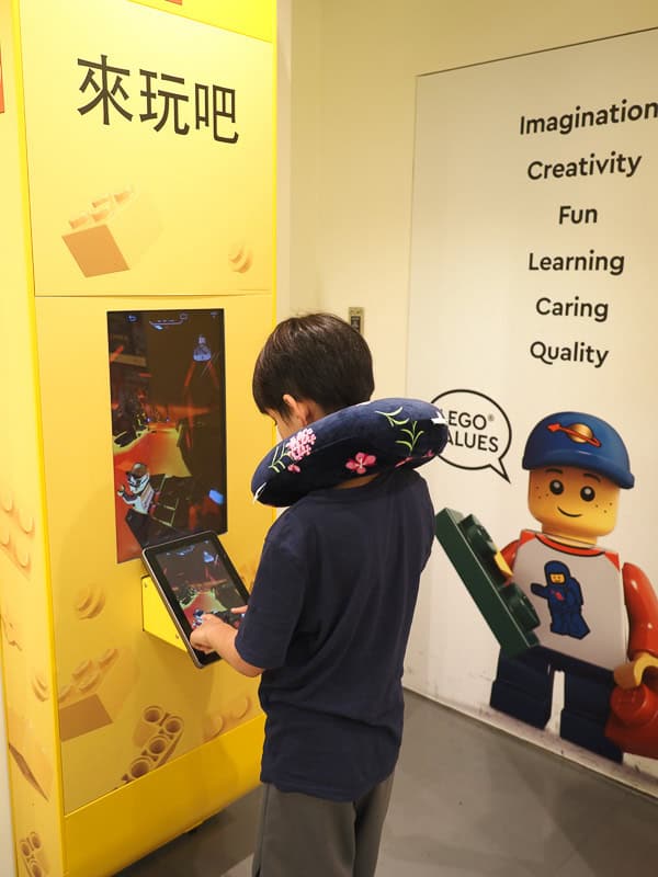 A young boy wearing airplane pillow around his neck playing a game on an iPad inside a LEGO store