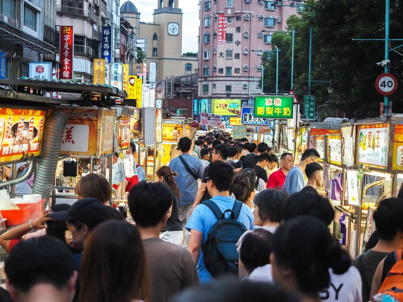Crowds of people in a narrow night market with food stalls and signs on either side