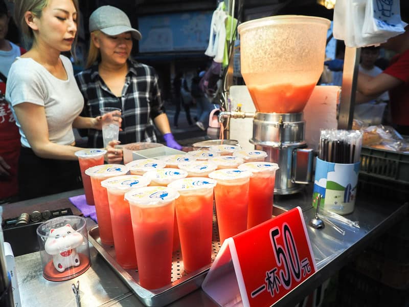 A night market vendor with about a dozen plastic cups of watermelon juice, juicer machine, and two women behind the stall preparing more