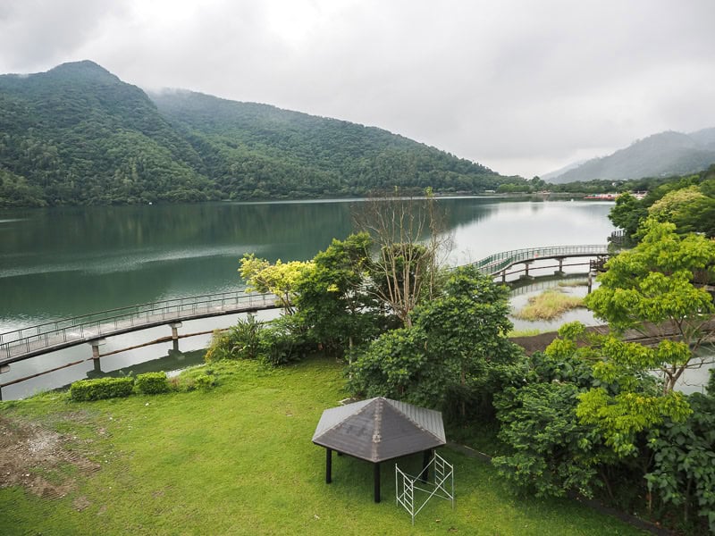 A green lawn in the foreground and raised wooden cycling path around a lake with mountains on opposite side