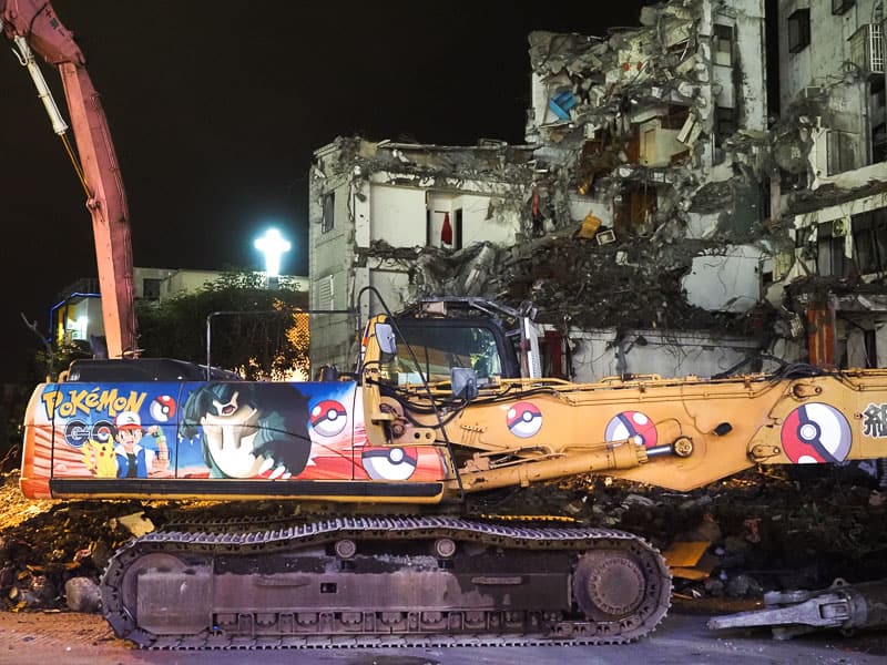 An orange excavator with Pokemon designs on it with a damaged building behind it, shot at night