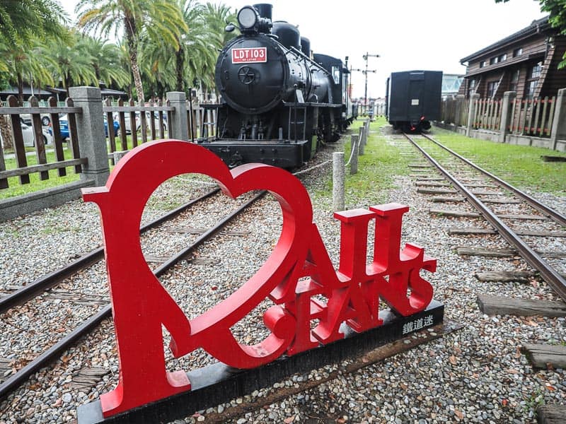 A red statue that spells the words I heart Railways sitting on a railway line with two black locomotives behind