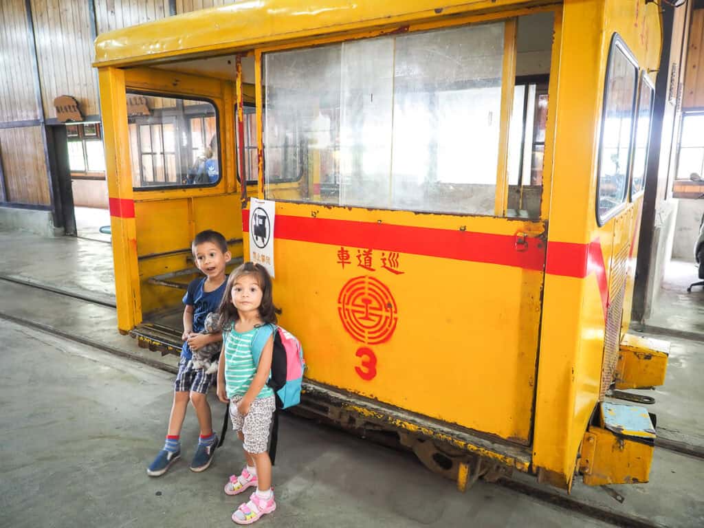 Two kids standing next to a small yellow train car inside a railway museum in Fenqihu