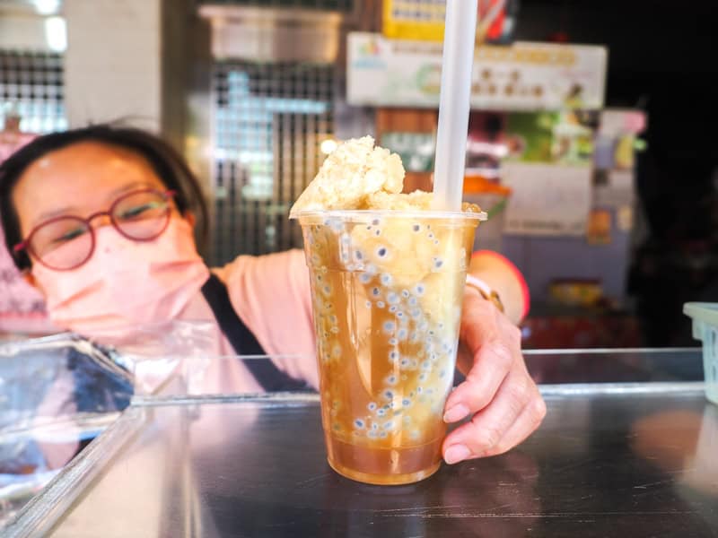 A Taiwanese vendor with mask passing a slushy drink with basil seeds across the glass counter to the camera