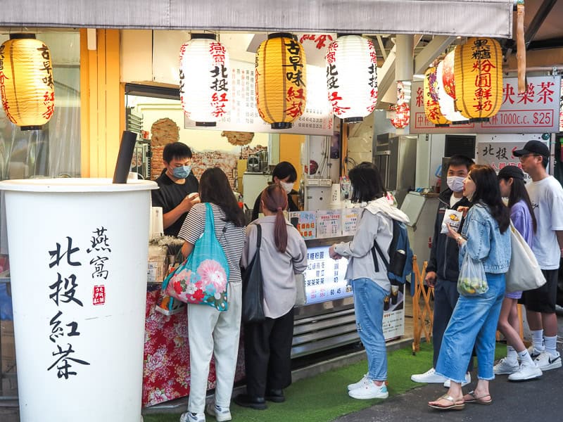 Several customers lined up and waiting for drinks in front of a Taiwanese iced tea shop, with several orange and white lanterns hanging above