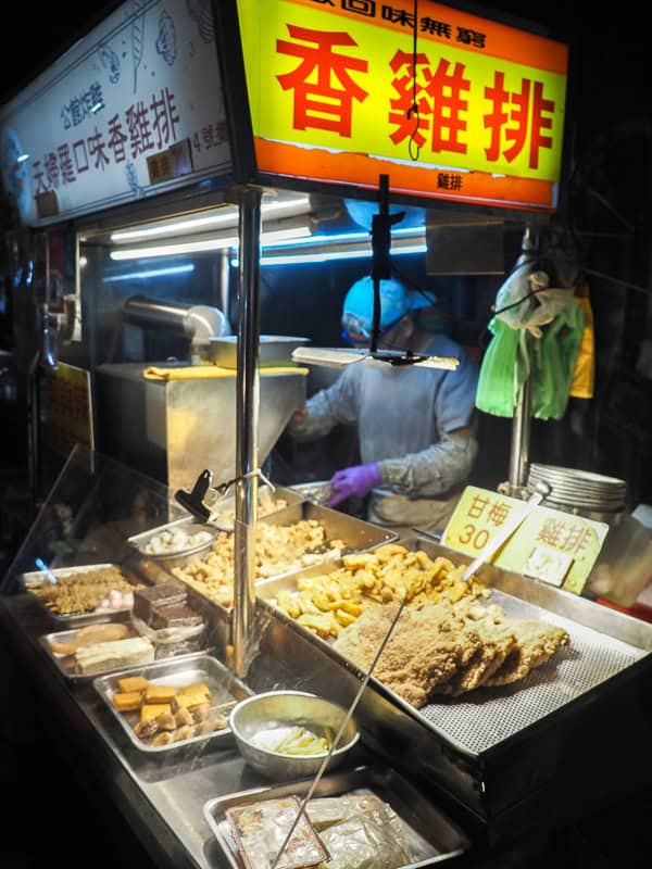 Vertical image of a night market stall with giant fried chicken fillets and other fried items on display and person behind preparing more