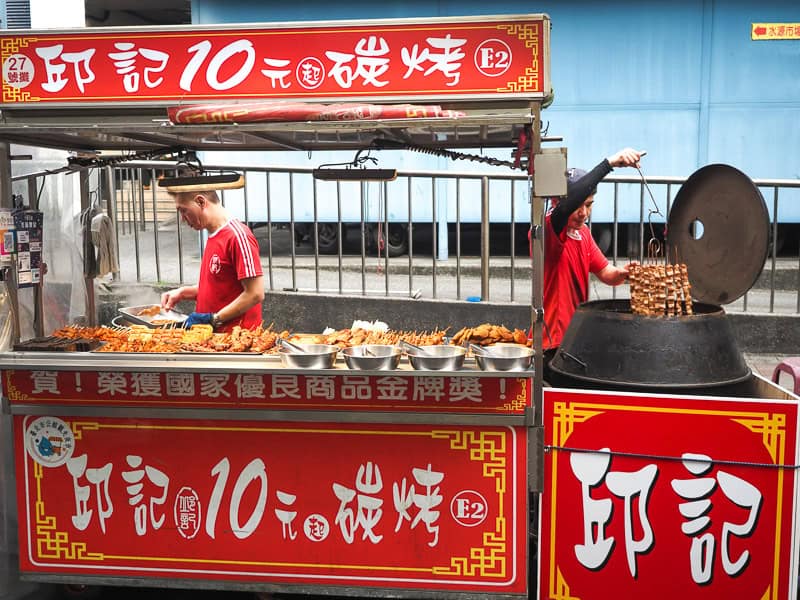 A night market food stall in the daytime, with red signs below and above it, many sticks of meat on display, a vendor working behind, and another vendor to the side dropping more beat sticks into a huge barrel oven