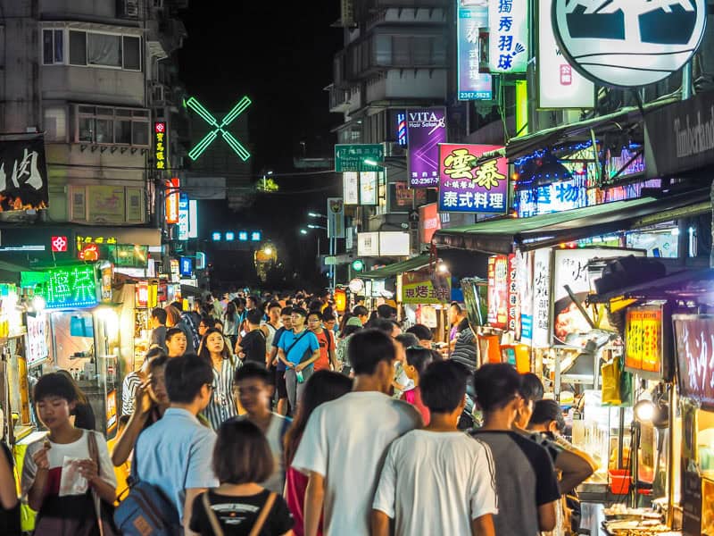 A colorful lively scene of many people walking through Gongguan Night market with lots of food stalls and colorful signs and lights on the sides